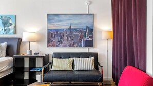 A modern hotel room with a black leather sofa, a cityscape painting, a lamp, and a red chair.