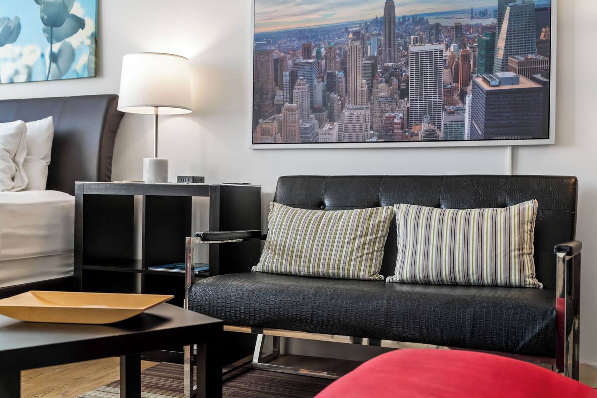 A modern hotel room with a black leather sofa, a white bed, a desk with a lamp, and a cityscape view.
