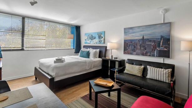 A modern hotel room with a large bed, a sofa, a desk, and a cityscape painting.