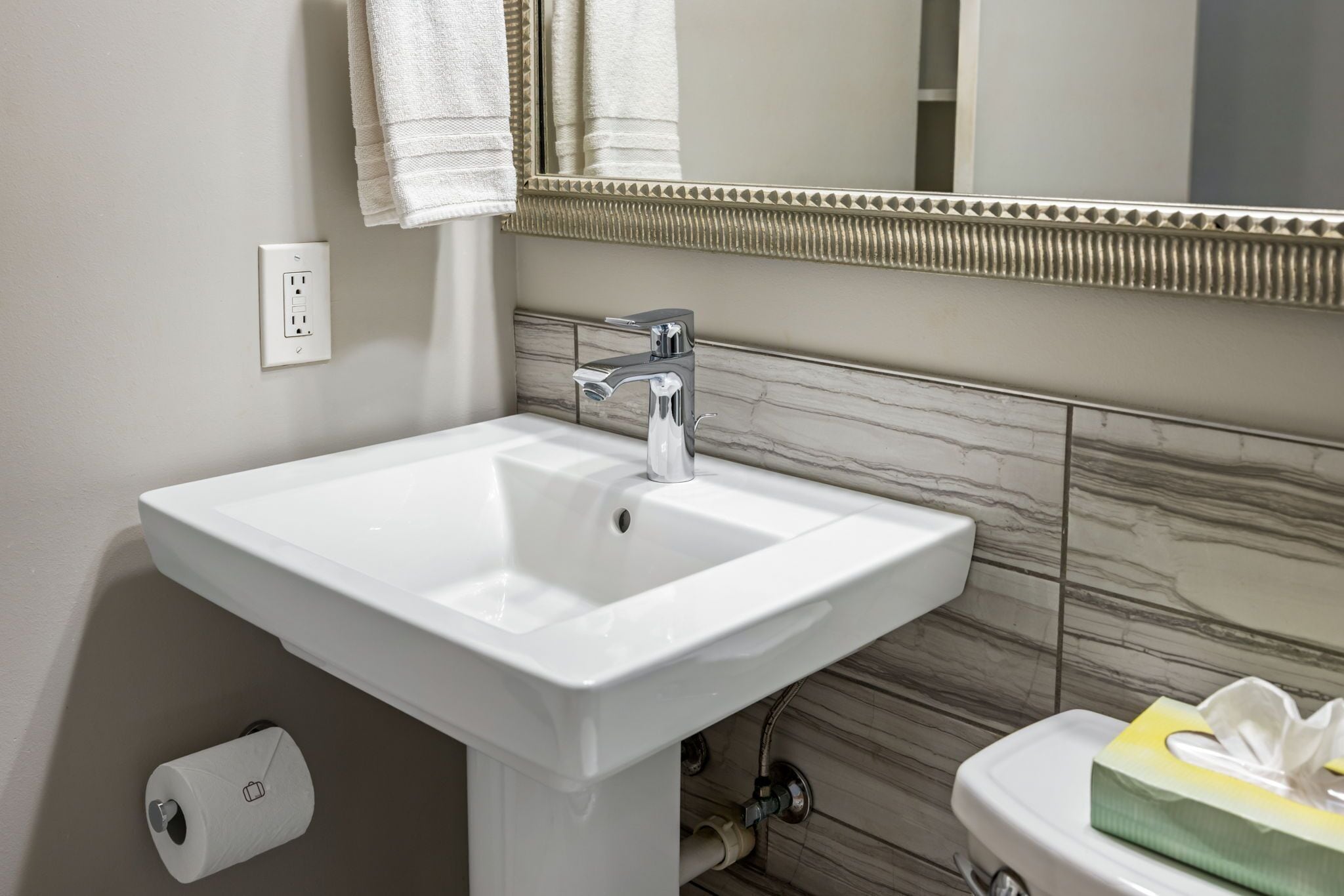 Une salle de bain avec un lavabo blanc, un miroir doté d’un cadre décoratif, un porte-serviettes et des toilettes avec un présentoir à mouchoirs.