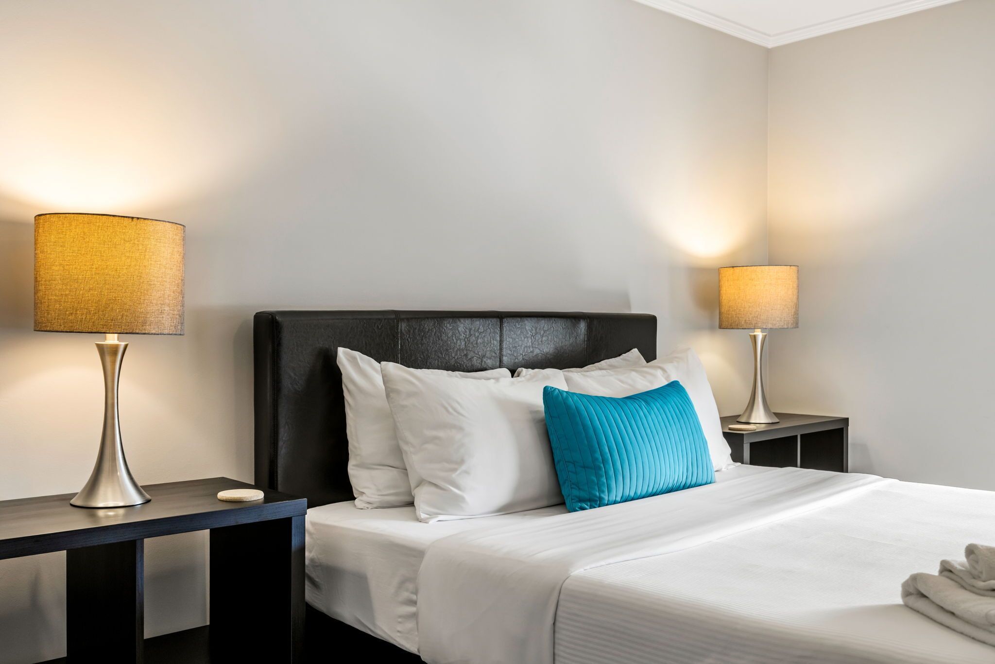 Un lit bien fait, avec une literie blanche et un coussin turquoise, flanqué de deux tables de chevet sur lesquelles sont posées des lampes.