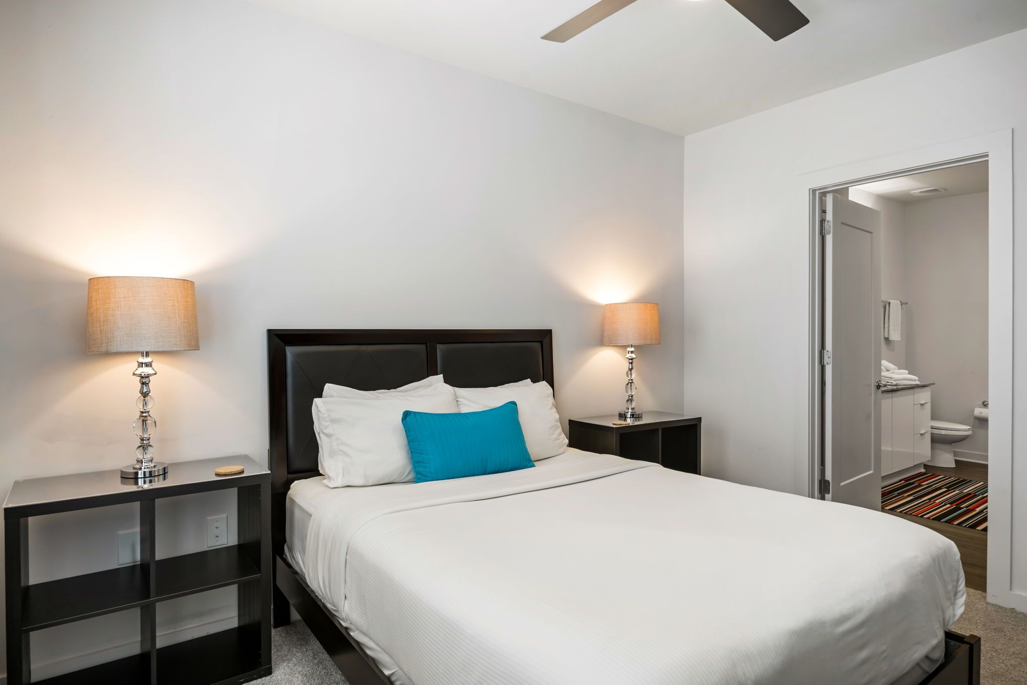A neatly arranged hotel room with a large bed, two bedside tables with lamps, a ceiling fan, and a view into the bathroom.