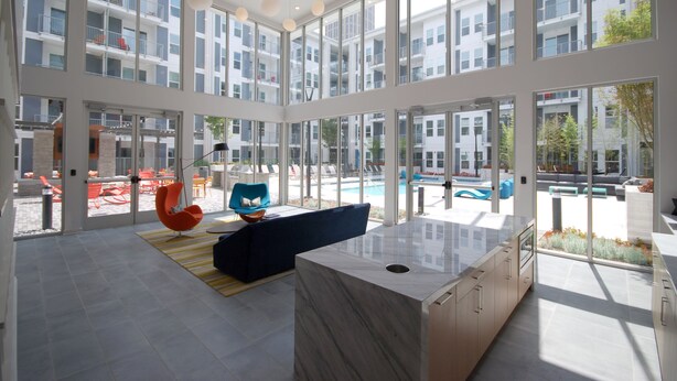 Un espacio interior amplio con ventanas grandes, una cocina moderna y una zona de estar con sofá azul y sillones naranja.