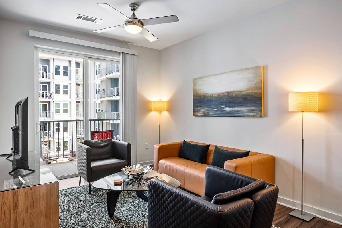 A modern living room with a sofa, armchairs, a coffee table, and a TV. There is a balcony with a view of buildings.