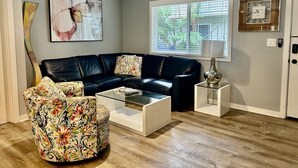 Una sala de estar con un sofá de cuero oscuro, una mesa de centro de vidrio, un reposapiés estampado con flores, un ventilador de techo, un ventanal con persianas y un reloj de pared.