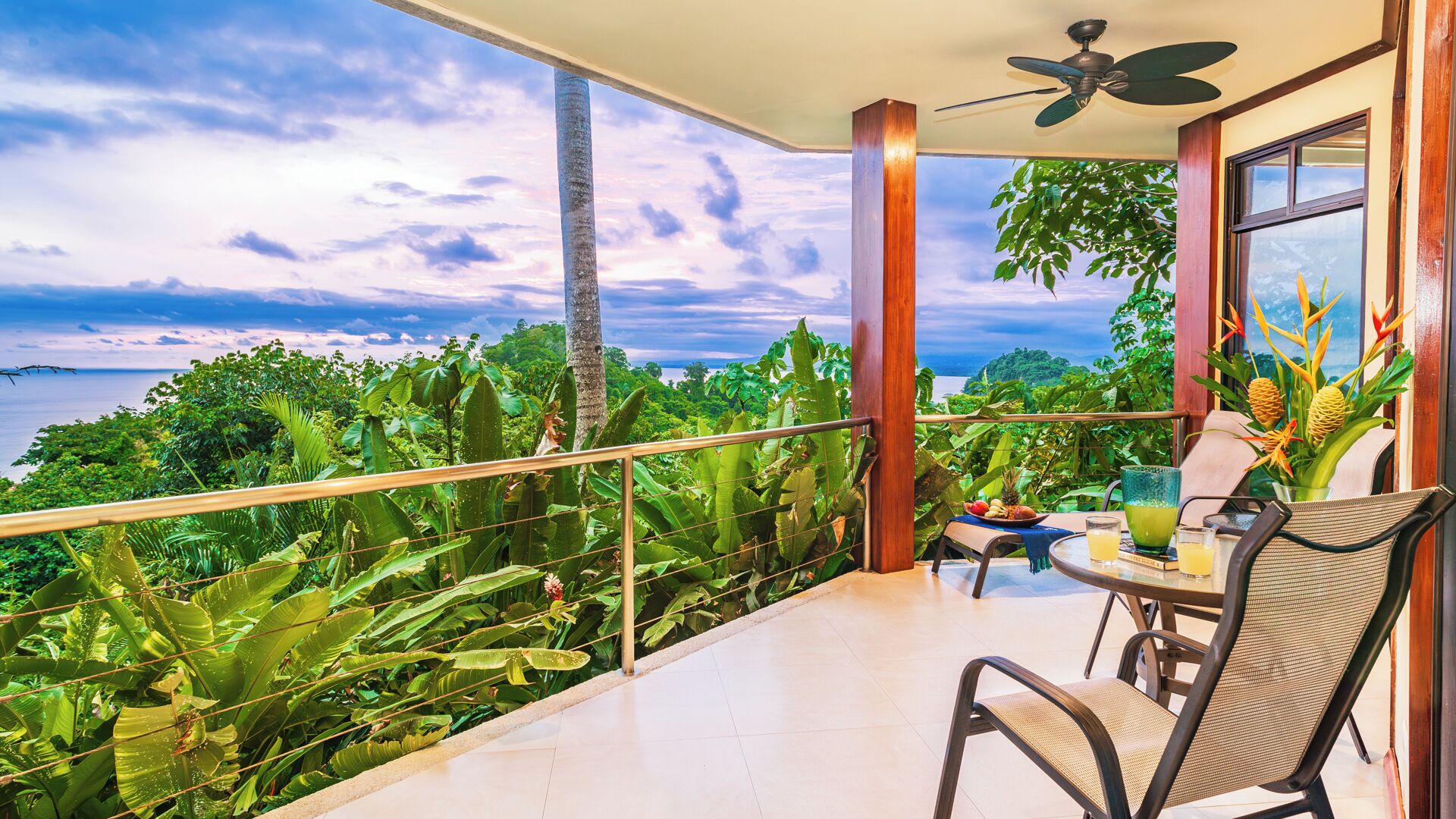 Un balcón con vistas a un paisaje tropical, una mesa con sillas y un ventilador de techo.