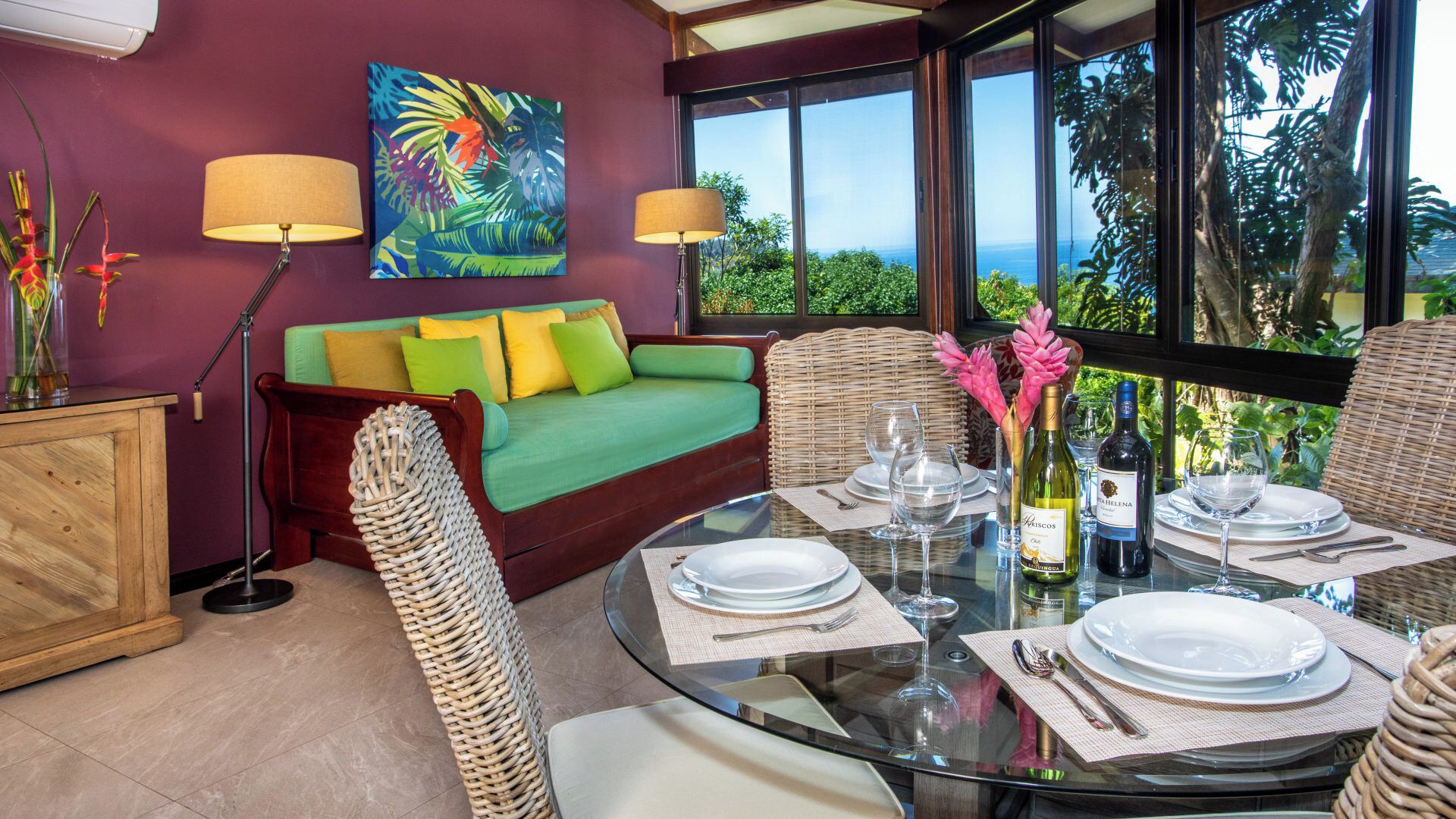 Un comedor con mesa de vidrio dispuesta para cuatro personas, un sofá verde, sillas de mimbre y vista al océano.