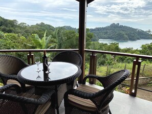 A round table with four wicker chairs, a bottle, and two wine glasses on a balcony overlooking a lush landscape and a body of water.