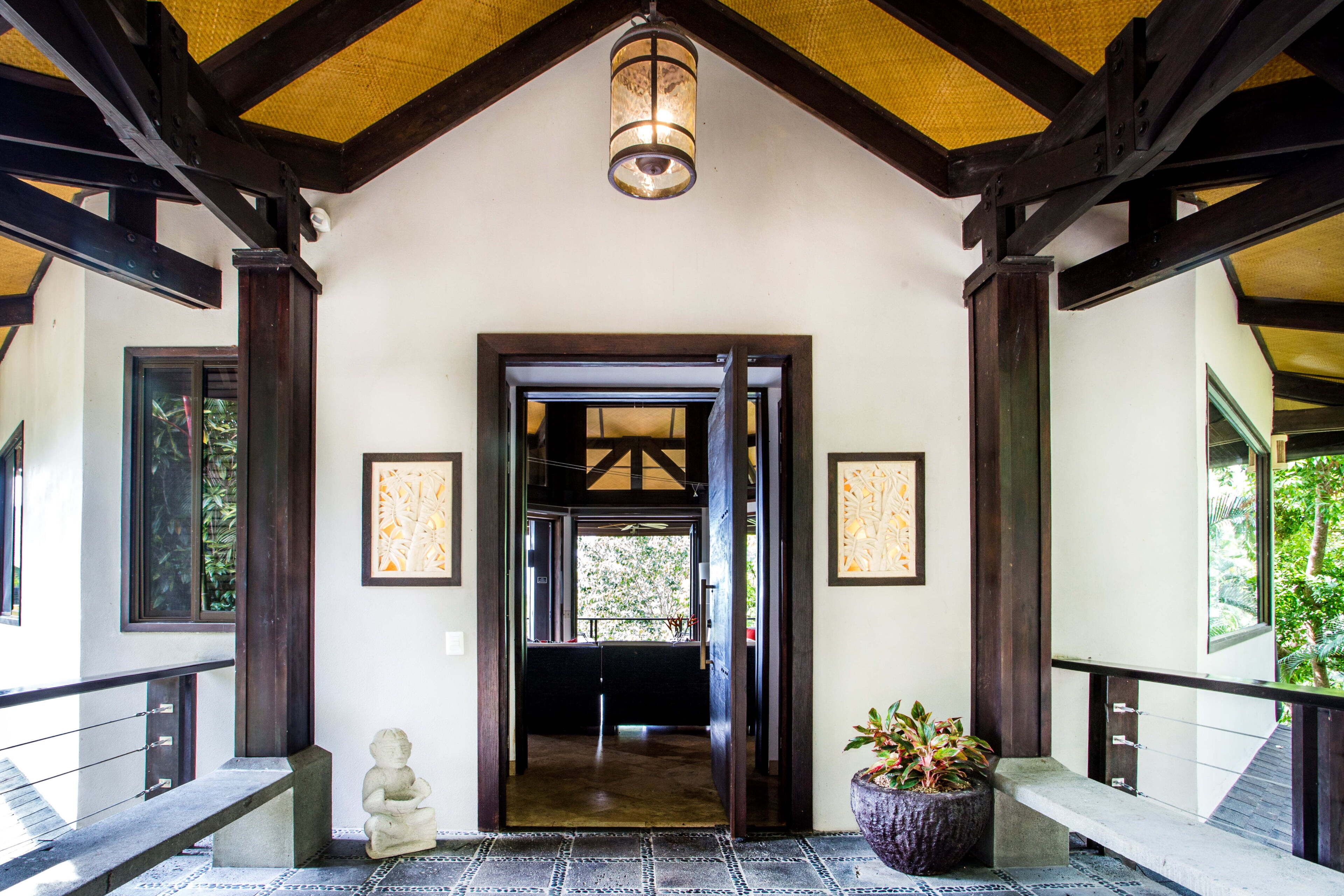 Entrada coberta com teto de madeira e luminária pendente. Há uma porta que dá para o interior, e um vaso de planta está posicionado no lado direito da entrada.