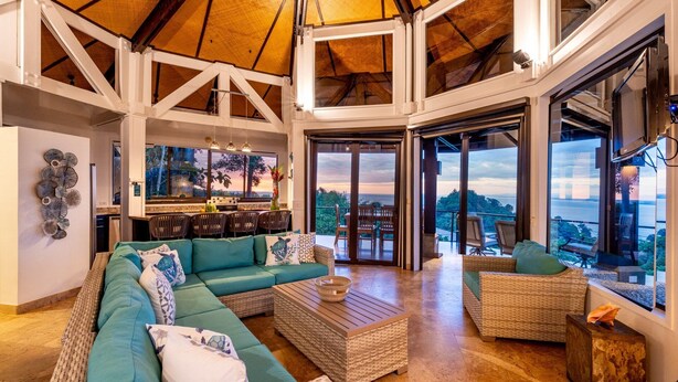 A spacious living area with a large sofa, a coffee table, and wicker chairs. There is a kitchen area with a bar and large windows offering a view of the outdoors. - Tulemar  (Quepos)