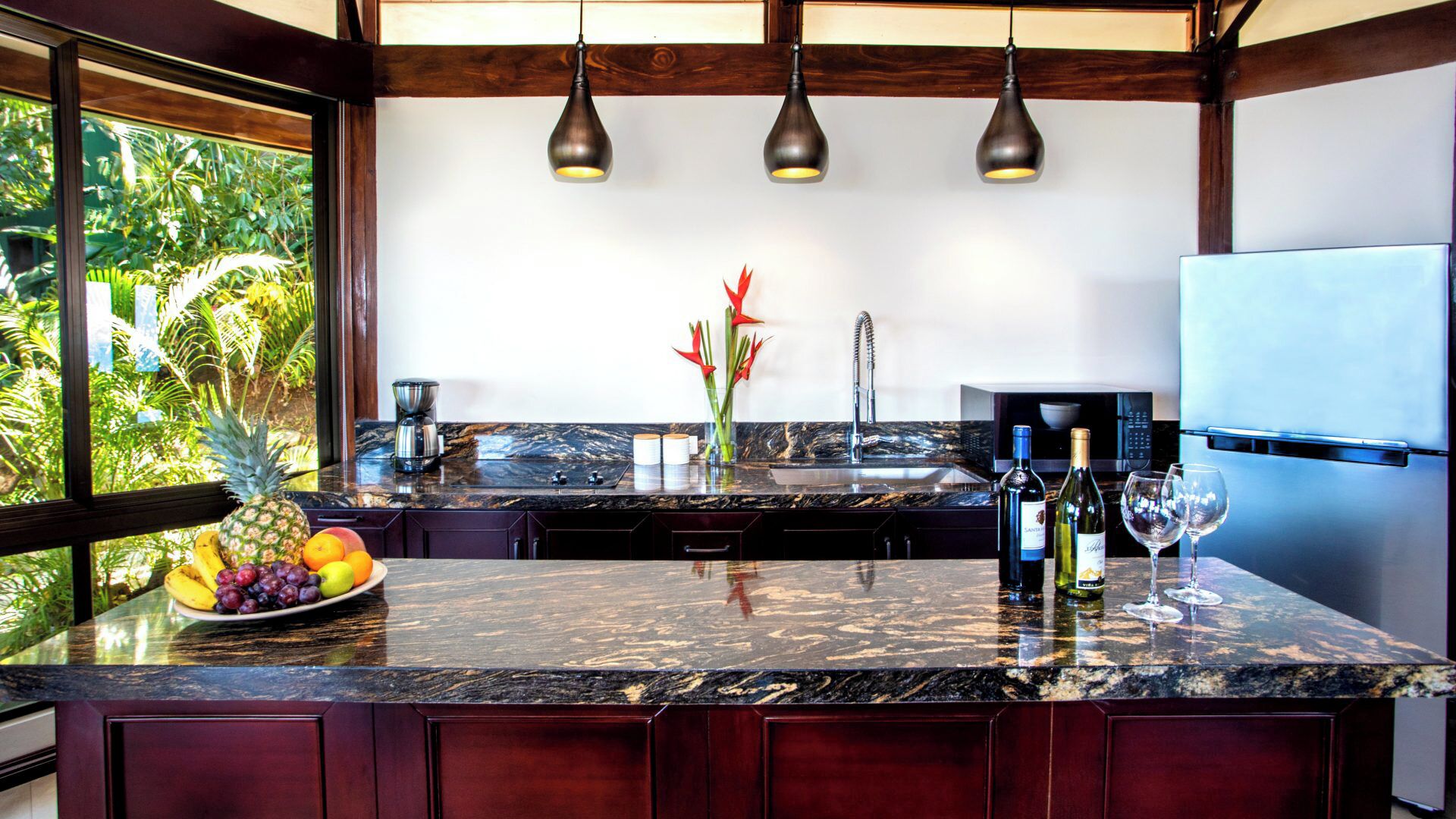 A modern kitchen with a granite countertop, a stainless steel refrigerator, and a window with a tropical view.