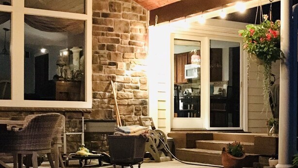 Un espacio al aire libre cubierto con paredes de piedra, techo de madera y un ventanal grande. Hay una silla, una mesa y una planta colgante.