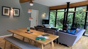 Una sala de estar moderna con una mesa de comedor de madera, sofá gris y un amplio ventanal con vistas al verdor.