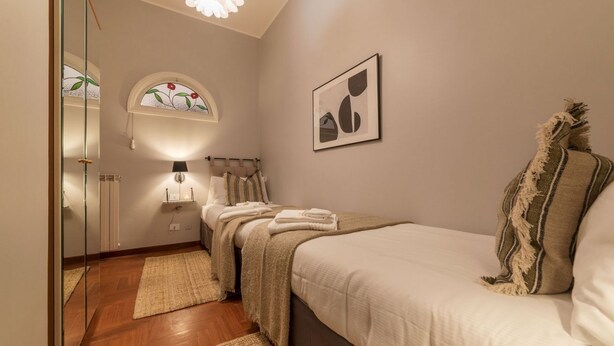 Un dormitorio con cama, mesita de noche, lámpara, espejo, ventana con diseño de vitral y un cuadro enmarcado en la pared.