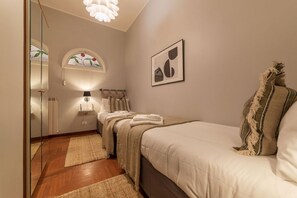 A bedroom with a bed, a bedside table, a lamp, a mirror, a window with a stained glass design, and a framed picture on the wall.