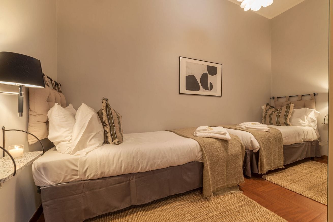 A hotel room with two beds, a bedside table with a lamp, a framed abstract artwork, and a woven rug.