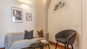Una sala moderna con un sofá gris, un sillón gris y una pequeña mesa redonda con un libro. Hay dos cuadros enmarcados en la pared y un ventanal decorativo con un diseño floral.