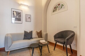 Ein modernes Wohnzimmer mit einem grauen Sofa, einem grauen Sessel und einem kleinen runden Tisch mit einem Buch. An der Wand hängen zwei gerahmte Bilder und das Fenster ist mit einem floralen Dekor verziert.