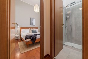 A bedroom with a bed, bedside table, and a nightstand. There is a shower area with a glass door and a towel rack.