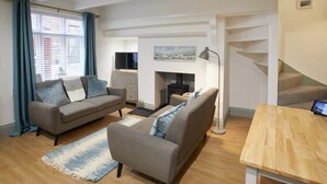 A modern living room with a sofa, armchair, and a small table. There is a fireplace and a staircase in the background. - Host & Stay | Threadneedle Cottage (Whitby)