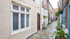 A narrow stone-paved alleyway between buildings with white and blue facades, a brown door, and potted plants. - Host & Stay | Threadneedle Cottage (Whitby)