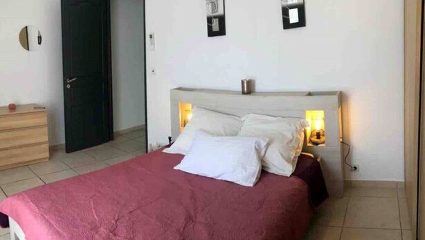 Ein ordentlich bezogenes Bett mit roter Tagesdecke und weißen Kissen, ein hölzerner Schminktisch und eine dunkle Tür.