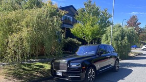 Un coche de lujo negro estacionado en una calle residencial con abundante vegetación y un edificio moderno al fondo.