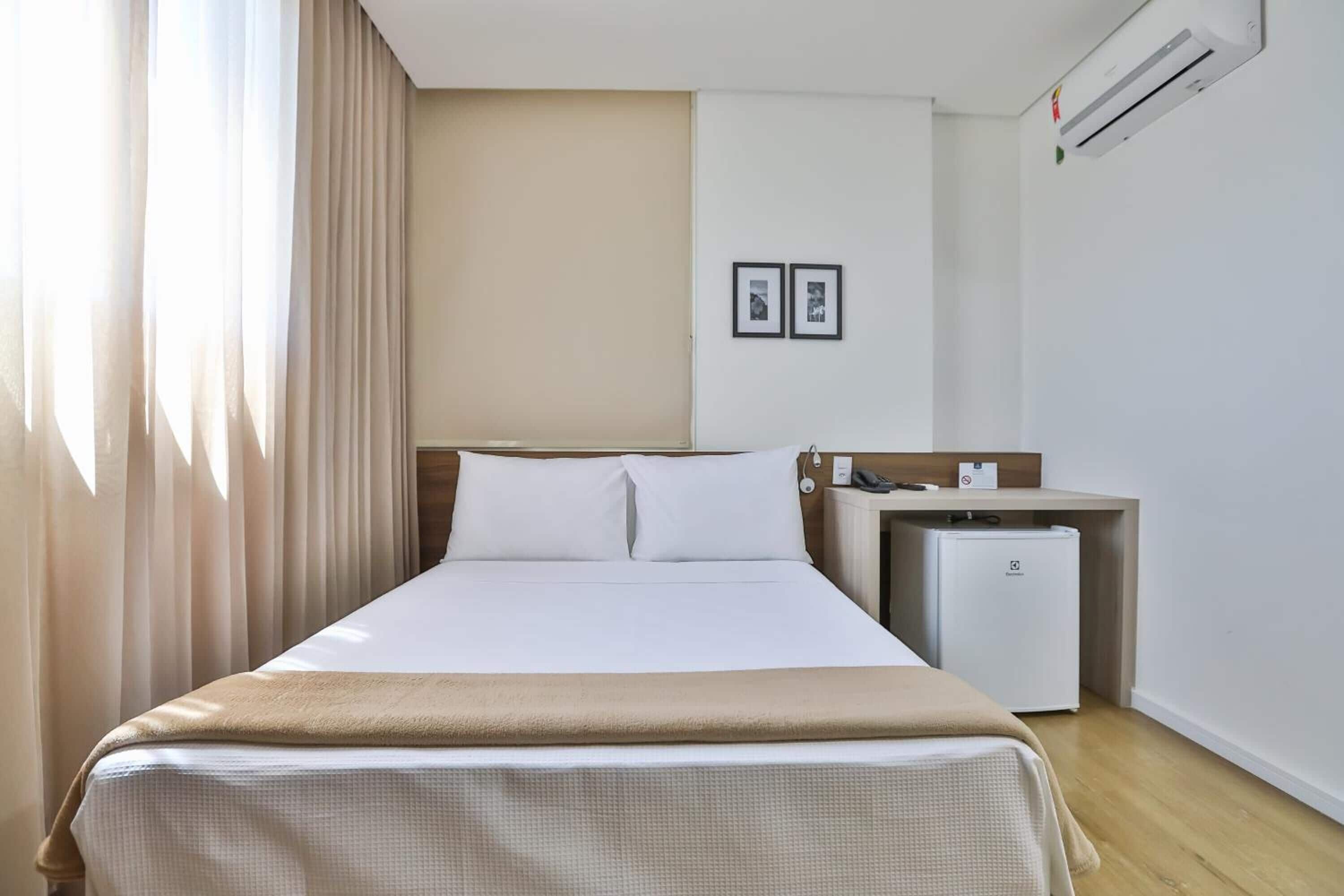 A hotel room with a bed, a small desk, a refrigerator, and a wall-mounted air conditioner.