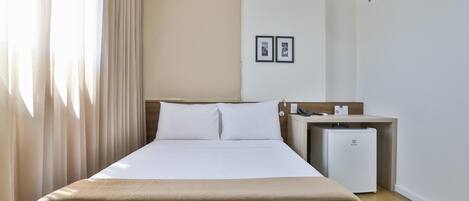 A hotel room with a bed, a small desk, a refrigerator, and a wall-mounted air conditioner.