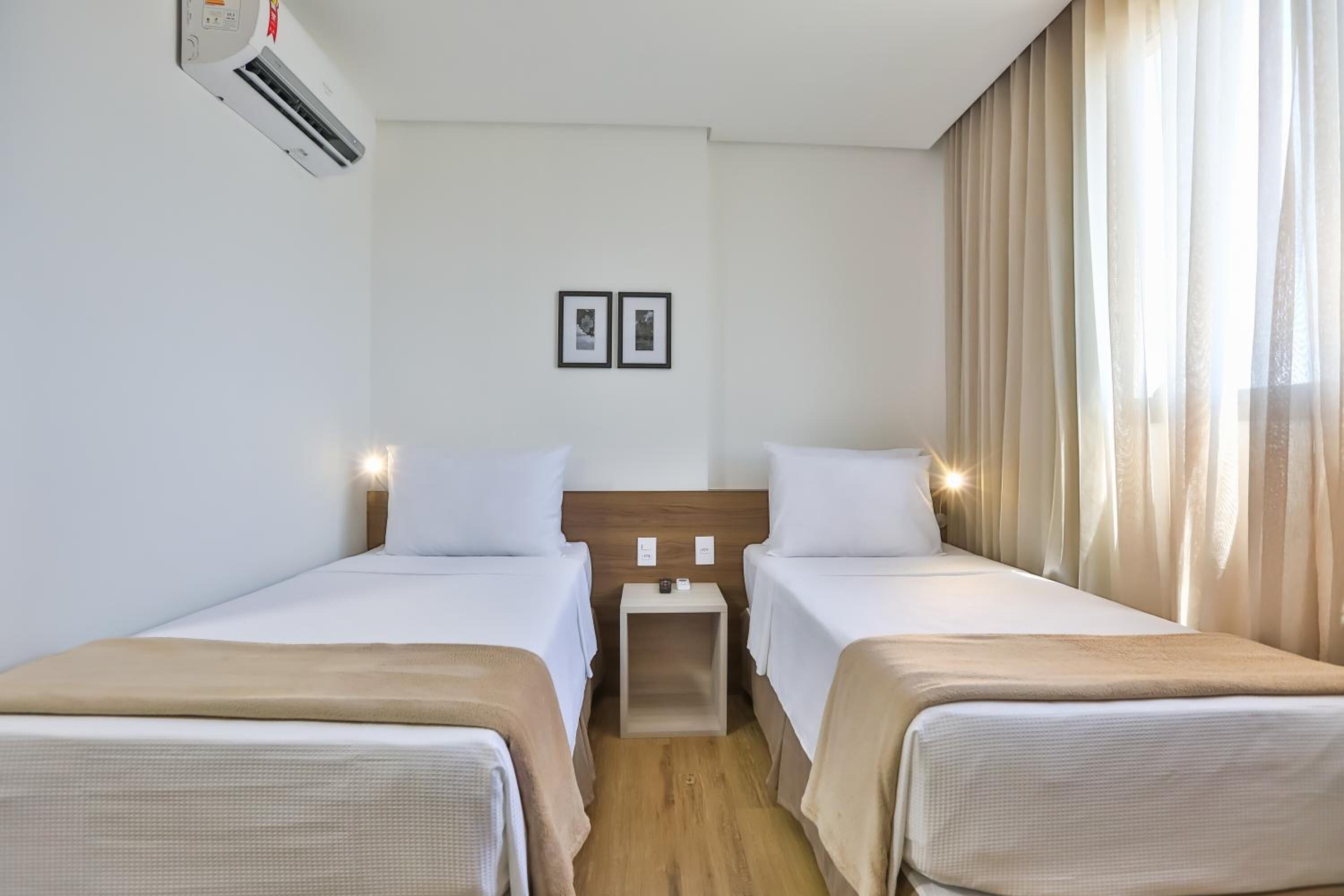 Una habitación de hotel con dos camas individuales, una mesita de noche pequeña, aire acondicionado en la pared y una ventana con cortinas.