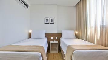 Una habitación de hotel con dos camas individuales, una mesita de noche pequeña, aire acondicionado en la pared y una ventana con cortinas.