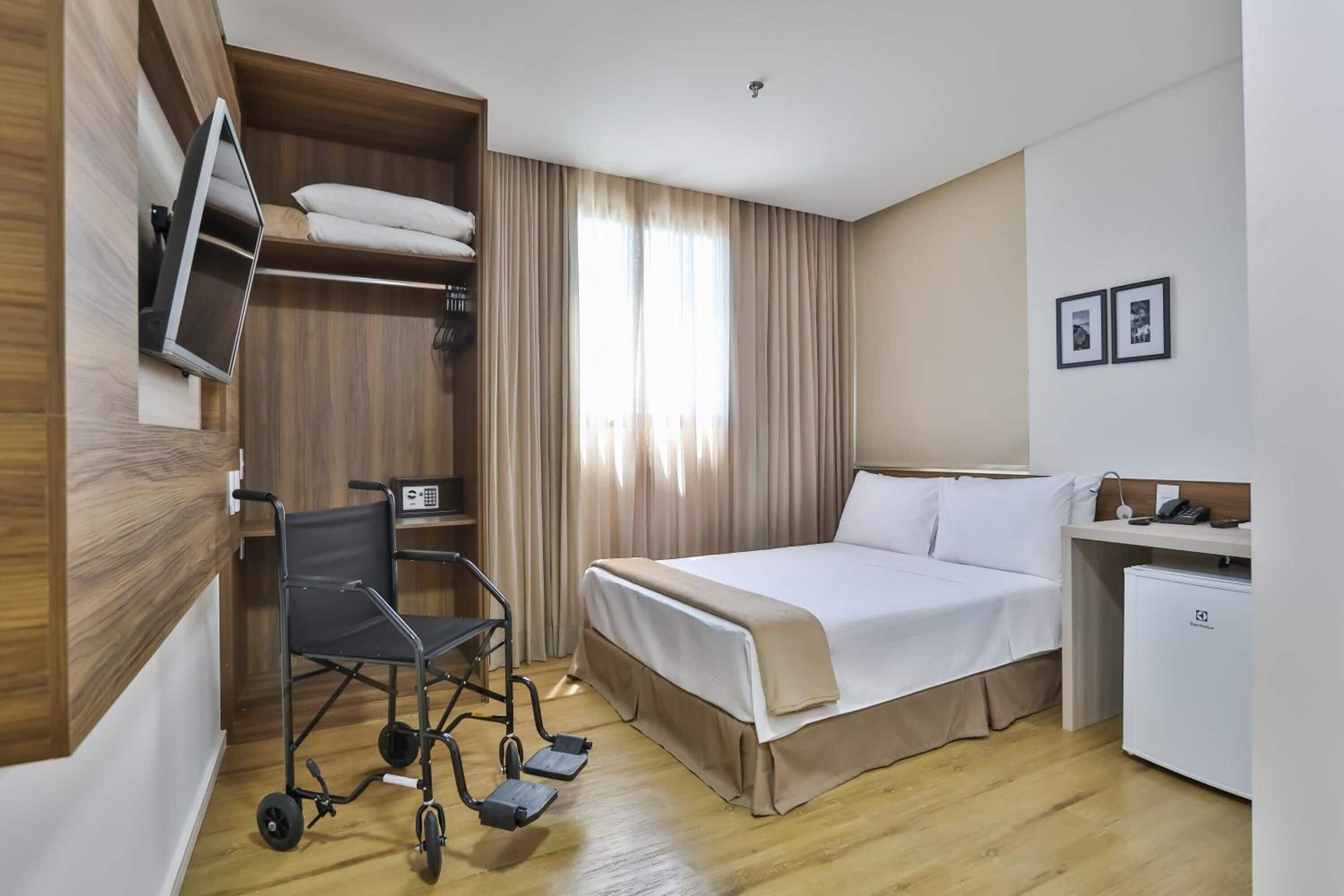 A hotel room with a bed, a desk, a chair, a TV, a mini-fridge, and a wheelchair.