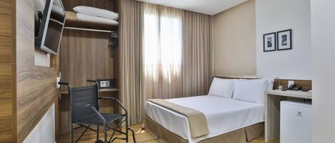 A hotel room with a bed, a desk, a chair, a TV, a mini-fridge, and a wheelchair.