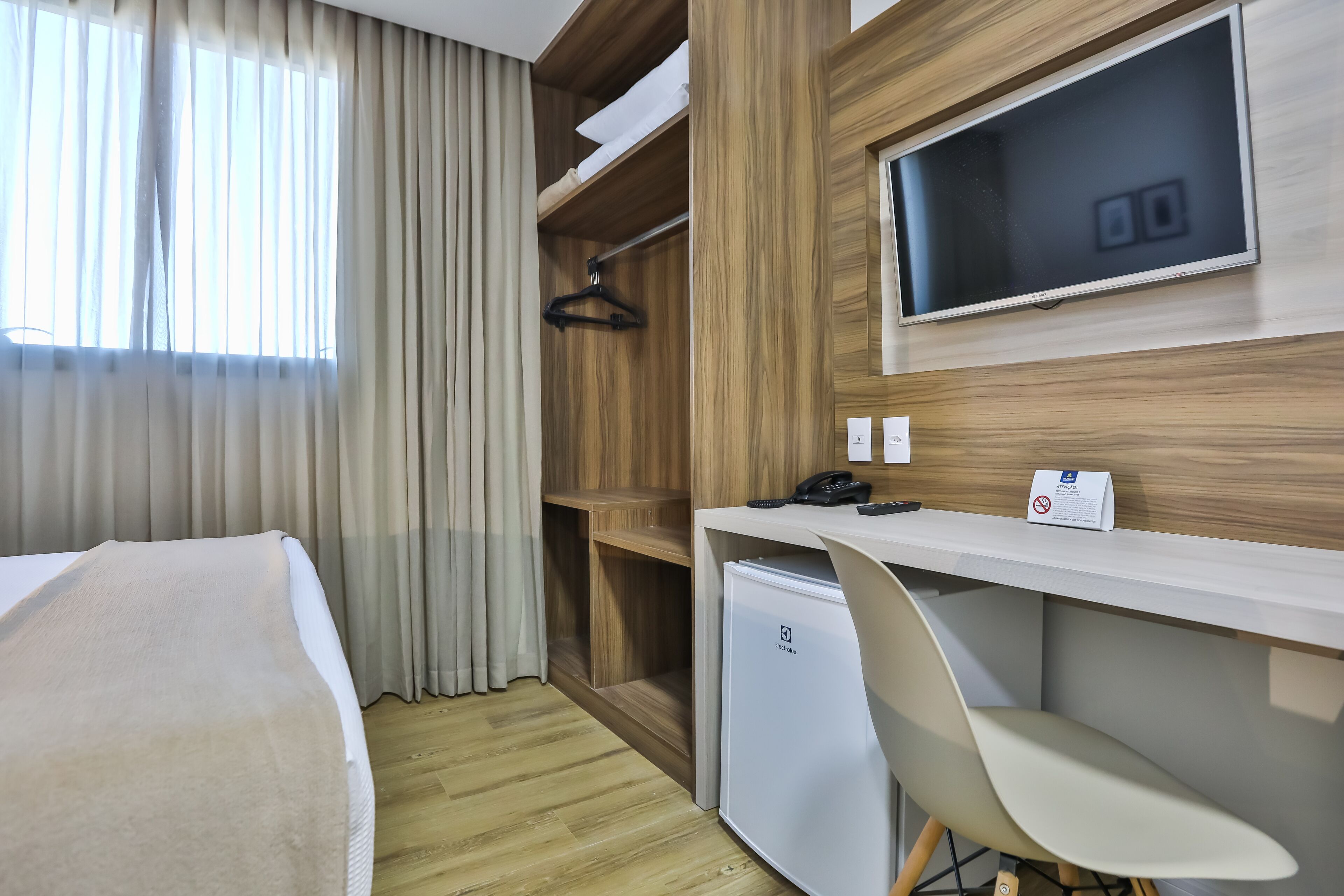 A hotel room with a bed, a desk, a chair, a small refrigerator, a TV, and a wardrobe.