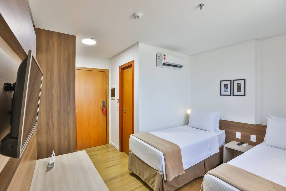 Ein Hotelzimmer mit zwei Betten, einem Fernseher, einem Holzschrank, einer Tür und einer Wandklimaanlage.