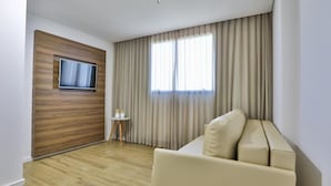 A modern hotel room with a wooden accent wall, a flat-screen TV, a beige sofa, and a large window with curtains. - Nobile Inn Meridional Cariacica (Cariacica)