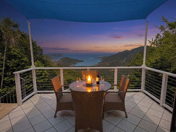 A covered outdoor seating area with a table and chairs, a candle, and a bottle, overlooking a scenic landscape.