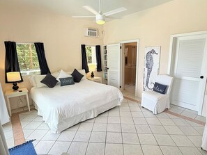A hotel room with a large bed, two bedside tables with lamps, a ceiling fan, a door leading to a bathroom, and a small chair with a cushion.