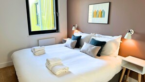 A neatly made bed with pillows, a bedside table, a wall-mounted lamp, and a window with blinds.