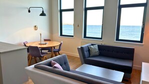 A modern living room with a sofa, a dining table with chairs, and large windows with a view of the sea.