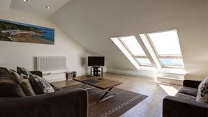 A spacious loft apartment with a high ceiling, a large window overlooking a beach, a cozy seating area with a sofa and armchairs, and a flat-screen TV on a stand. - Stables 2 -  a llyn peninsula that sleeps 2 guests  in 1 bedroom (Pwllheli)