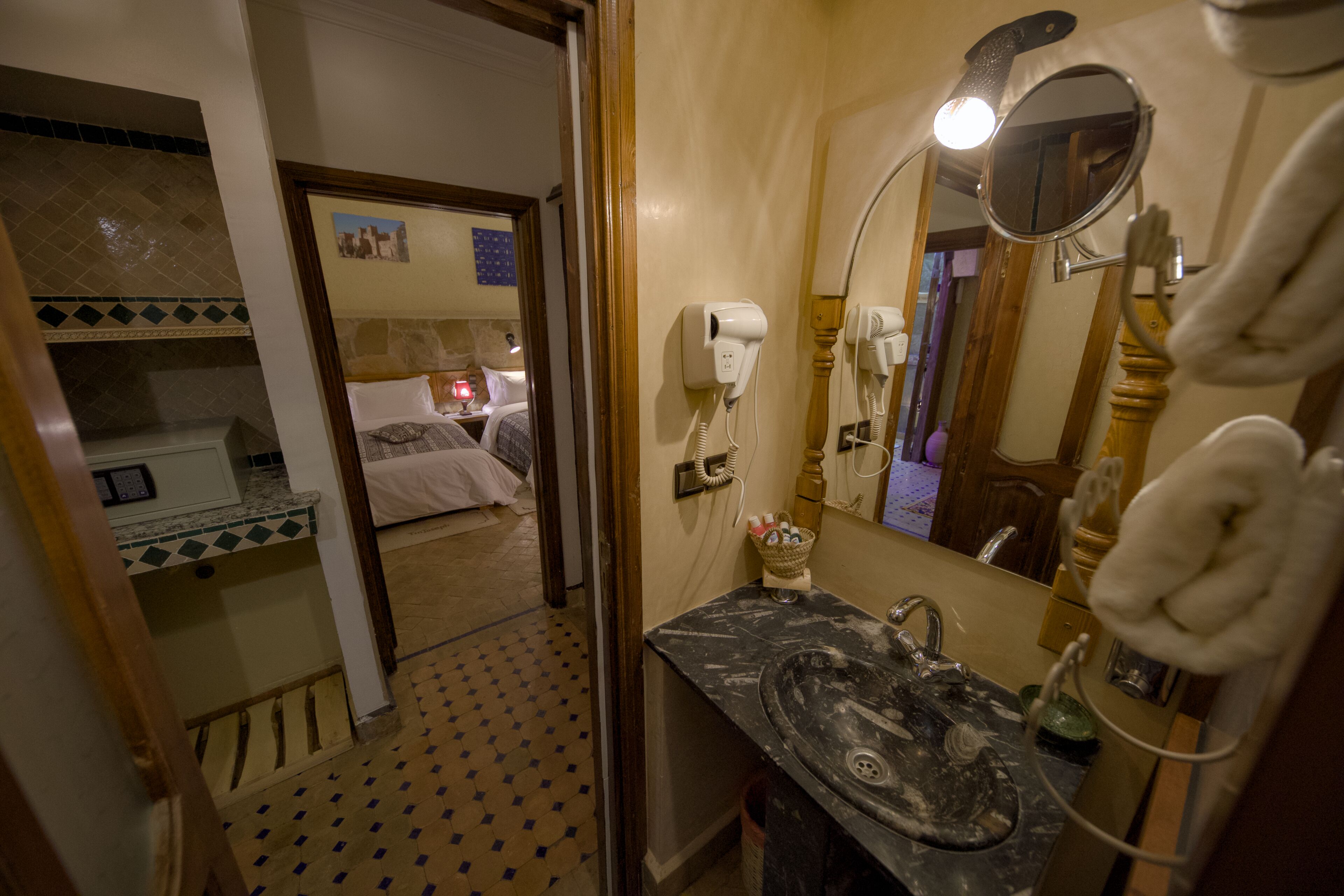 A bathroom with a sink, mirror, and hairdryer. There is a bed visible in the adjacent room.