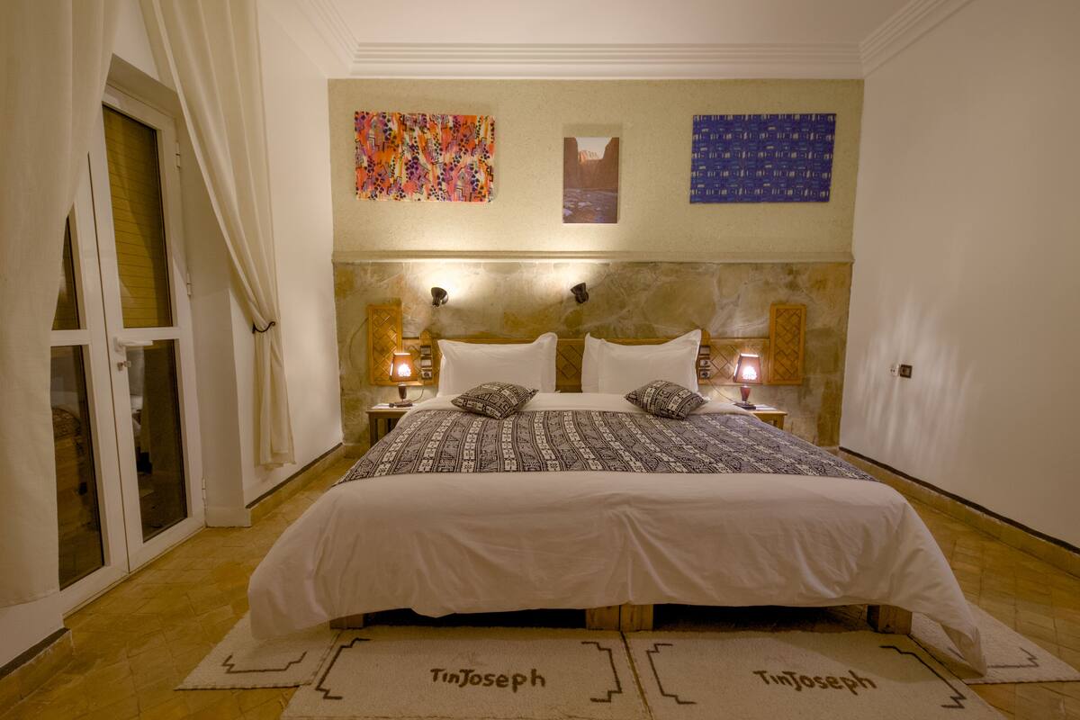 A bedroom with a large bed, two bedside lamps, a door with curtains, and a wall with two artworks.