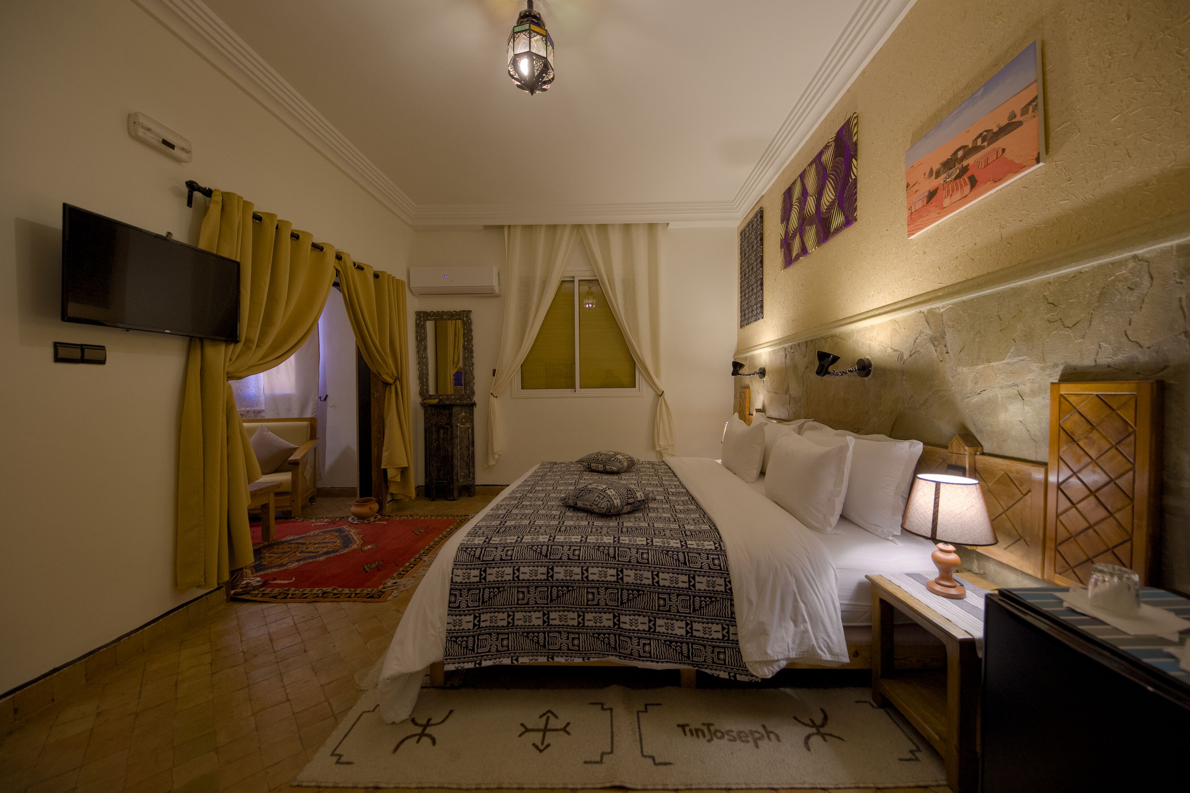 A hotel room with a bed, a TV, a lamp, a wall-mounted TV, and a window with curtains.