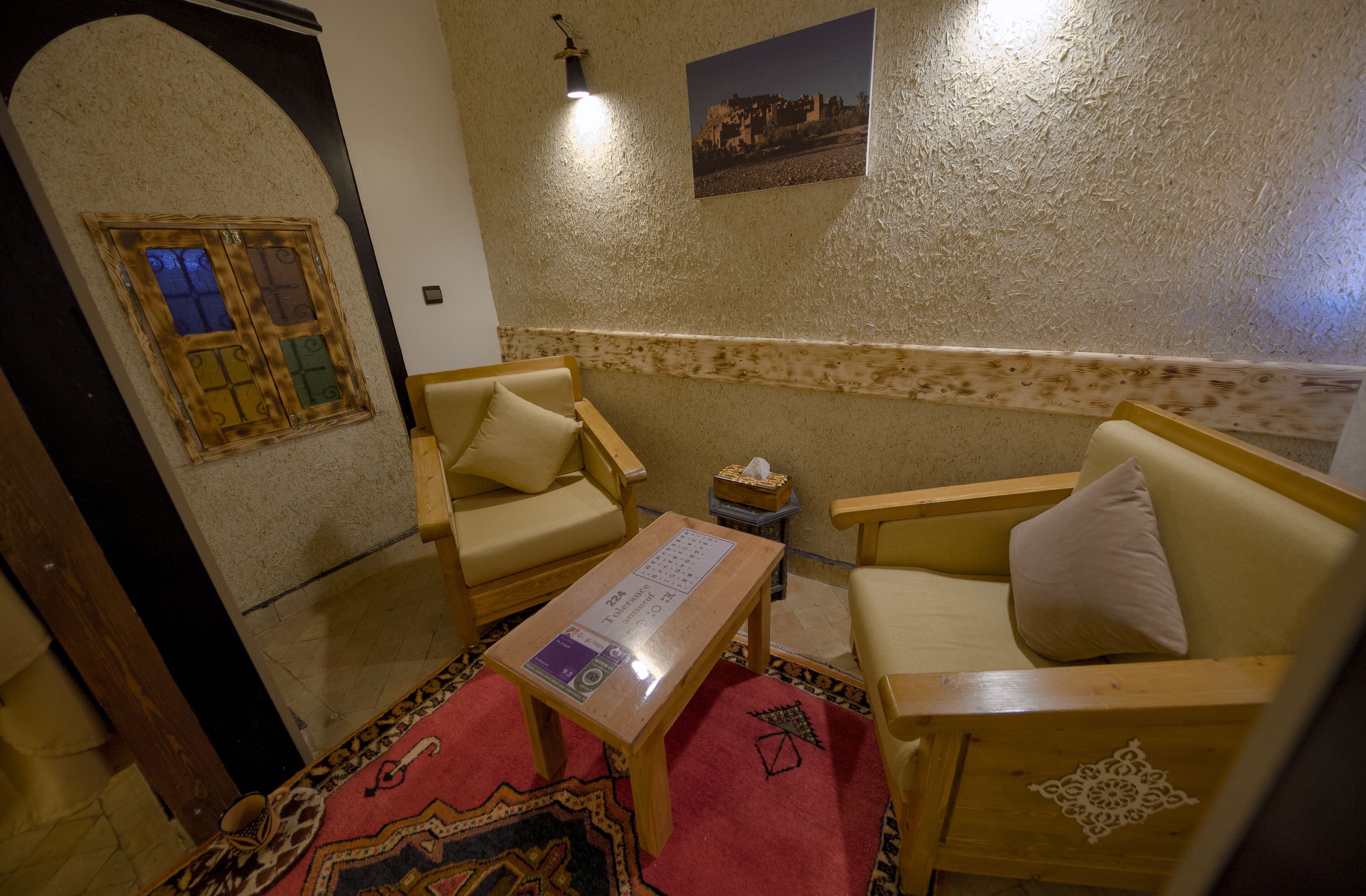 A cozy room with two wooden armchairs, a small table with a tissue box, a patterned rug, and a wall-mounted picture.