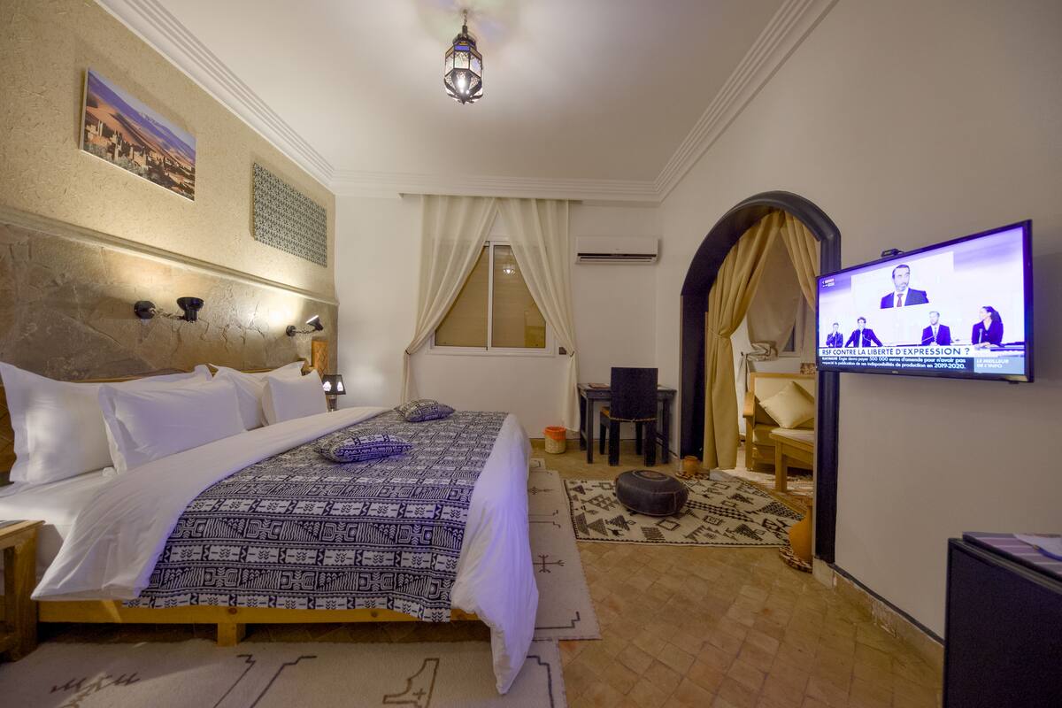 A hotel room with a large bed, a television displaying a news broadcast, a small table with a chair, and a decorative wall piece.