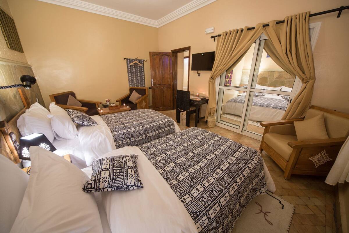 A bedroom with two beds, a desk, a chair, and a sliding glass door leading to a balcony.