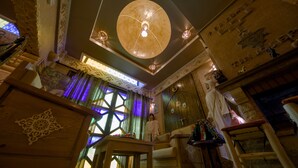 A cozy room with a large circular ceiling light, a wooden table, and a window with a geometric pattern. - Tin Joseph (Ouarzazate)