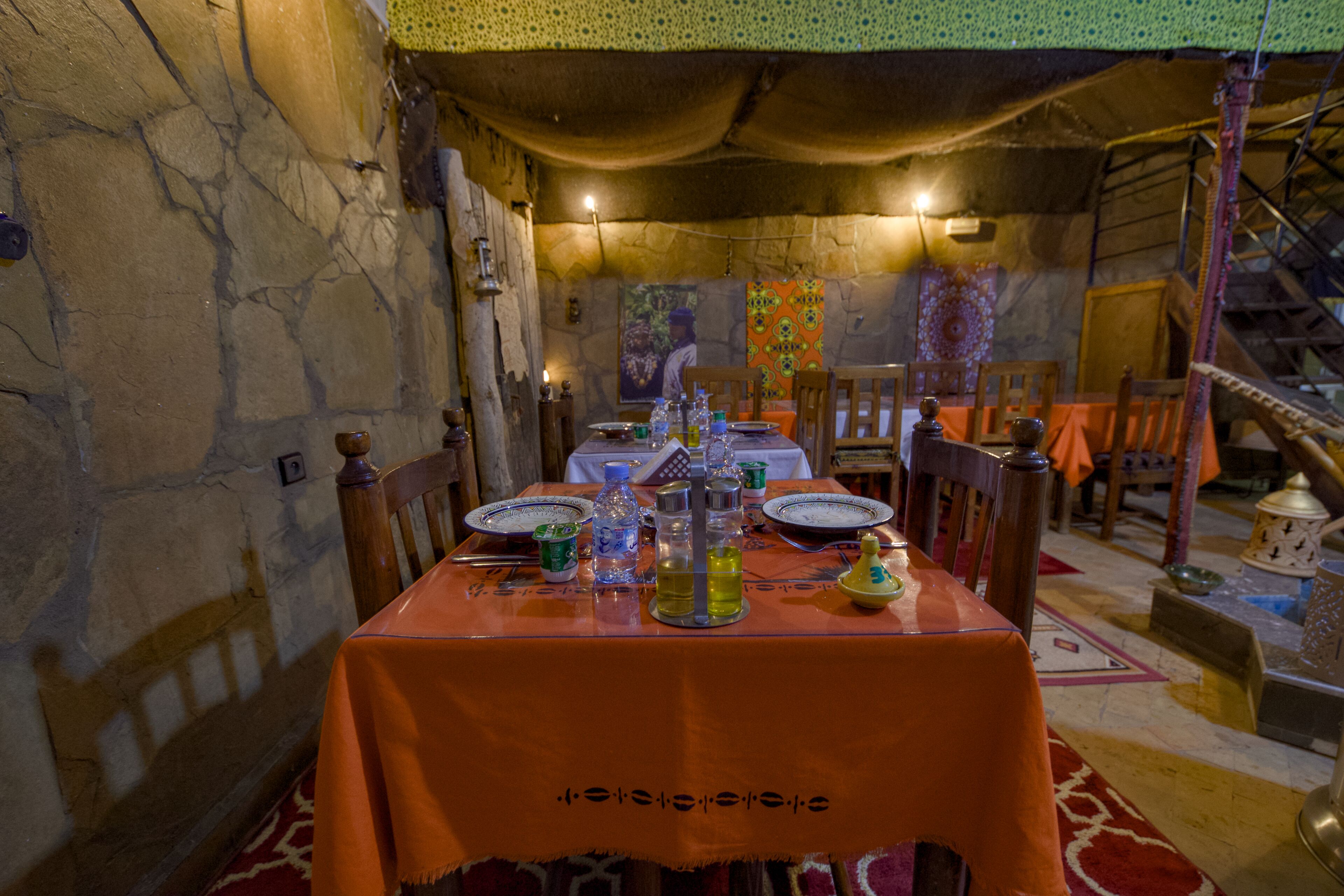 Un coin repas avec une table dressée pour deux personnes, agrémentée d’une nappe traditionnelle, d’assiettes, de verres et d’une bouteille. La pièce est dotée de murs en pierre et un escalier se trouve au fond.