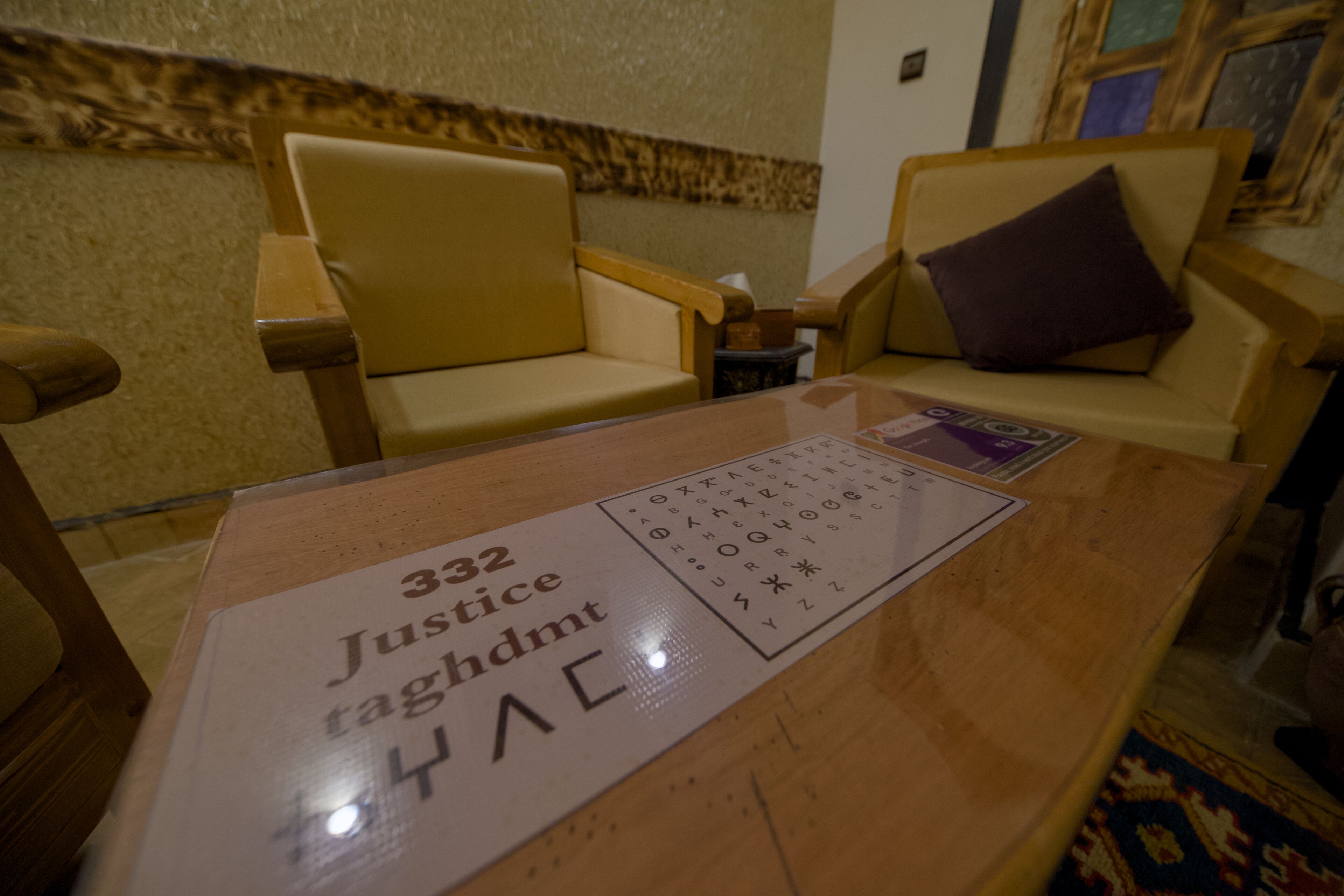 A room with a wooden table, two beige armchairs, and a small table with a sign reading "332 Justice taghdmt".
